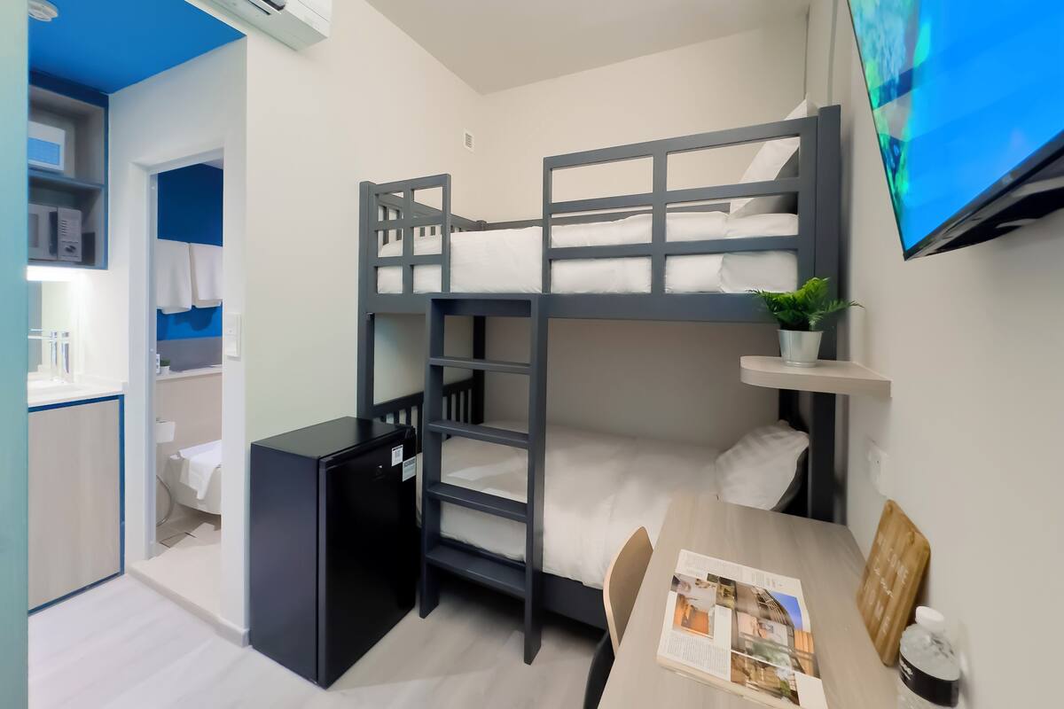 Een slaapkamer met een stapelbed, bureau, kleine koelkast, televisie en een plant.