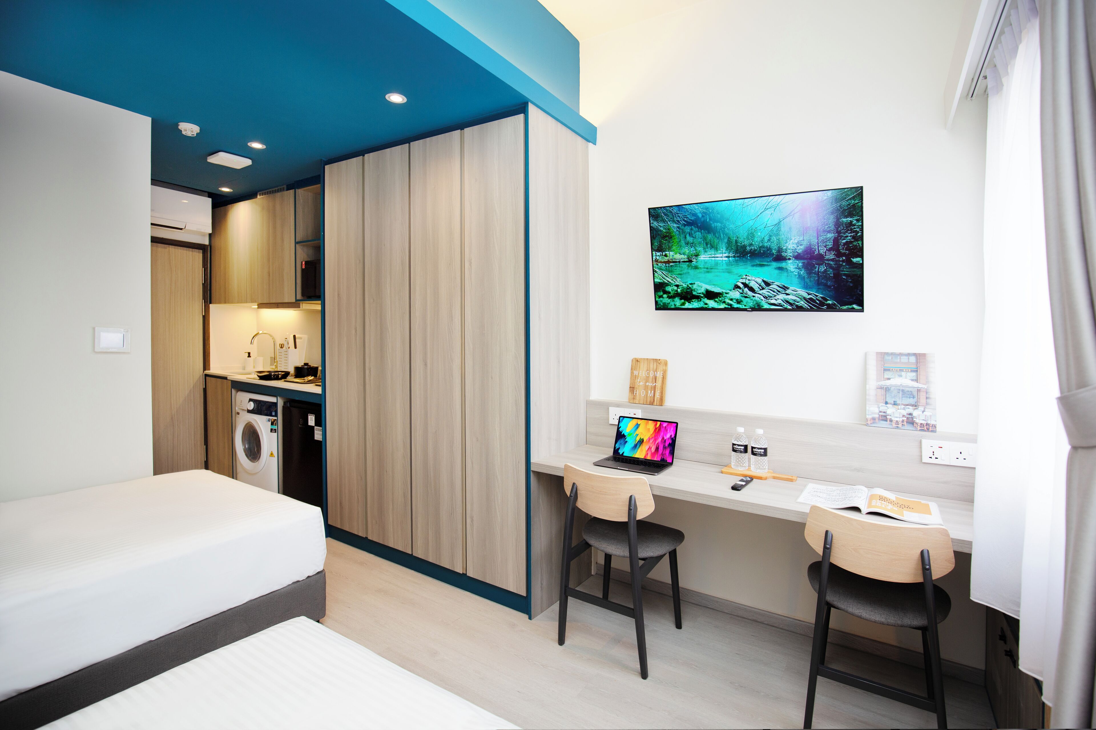 A modern hotel room with a bed, a desk with a laptop, a TV, and a washing machine.
