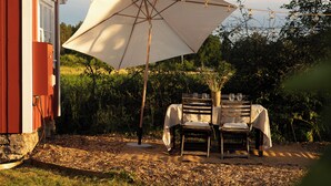 An outdoor dining area with a table and chairs under a large umbrella, a red building in the background, and a garden setting. - Cozy vacation home on Öland, close to nature and the sea (Kalmar län)