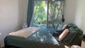 Ein Schlafzimmer mit einem Bett, einem Fenster mit Blick auf Grünflächen, einem Ventilator und einem Nachttisch mit Taschentuchbox.
