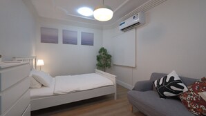 A neatly arranged bedroom with a single bed, a bedside table with a lamp, a grey sofa with patterned cushions, and a wall-mounted air conditioner. - 2-bedroom apartment IKEA style (Riyadh)