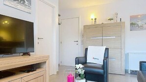 A living room with a flat-screen TV, wooden entertainment unit, a black leather chair, a glass vase with flowers, and a cabinet with a towel on it. - Zandloper III with terrace by Interhome (Bredene)