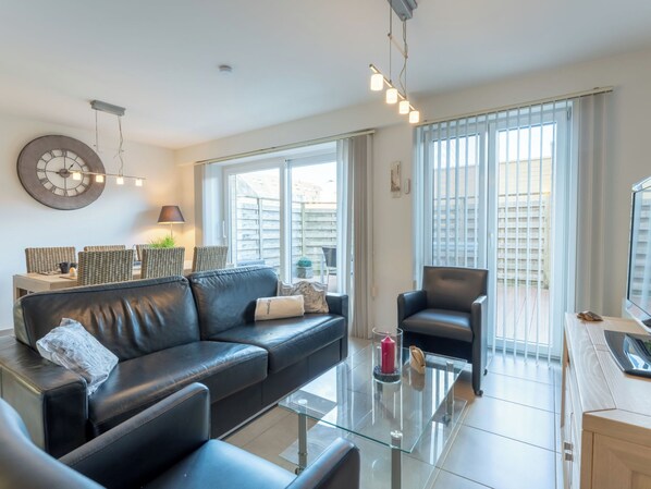 A modern living room with a black leather sofa, a glass coffee table, and a dining area with wicker chairs.