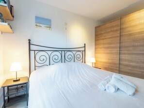 A neatly made bed with white linens and two rolled towels, a bedside table with a lamp, a bookshelf with books, and a wooden panel wall.