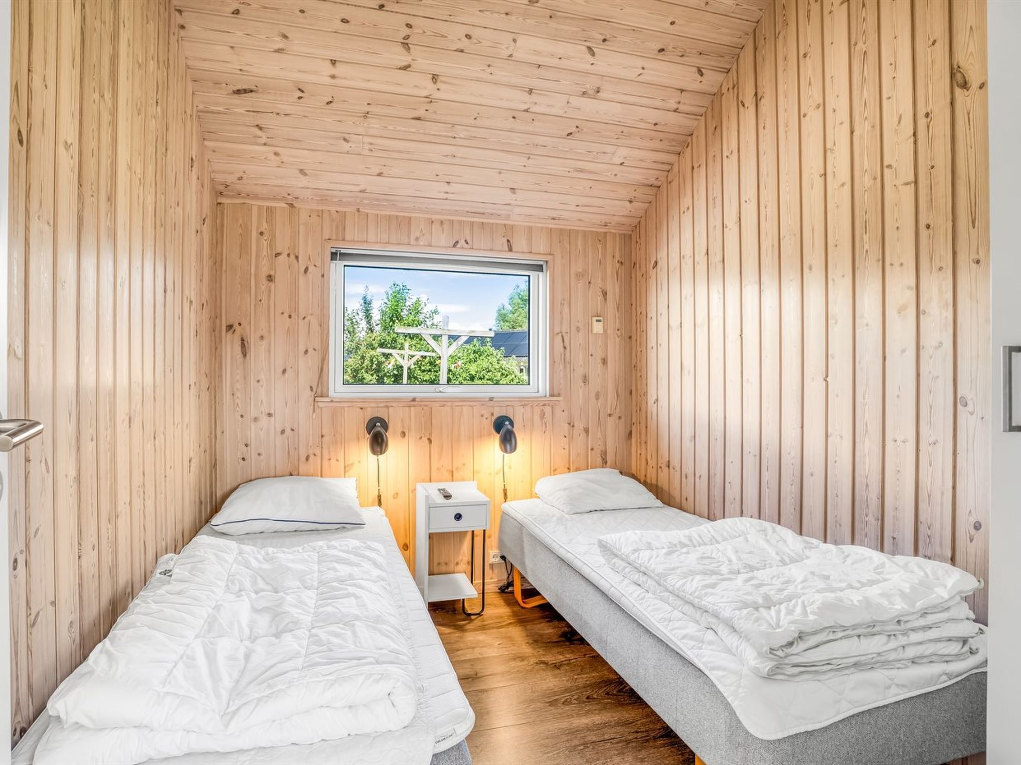 Ein Zimmer mit Holzwänden und einem Fenster, zwei Betten, Nachttischen und Blick auf Grün.