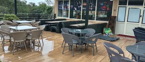 An outdoor patio area with wooden flooring, wicker tables and chairs, and a covered seating area with a view of the surrounding greenery.