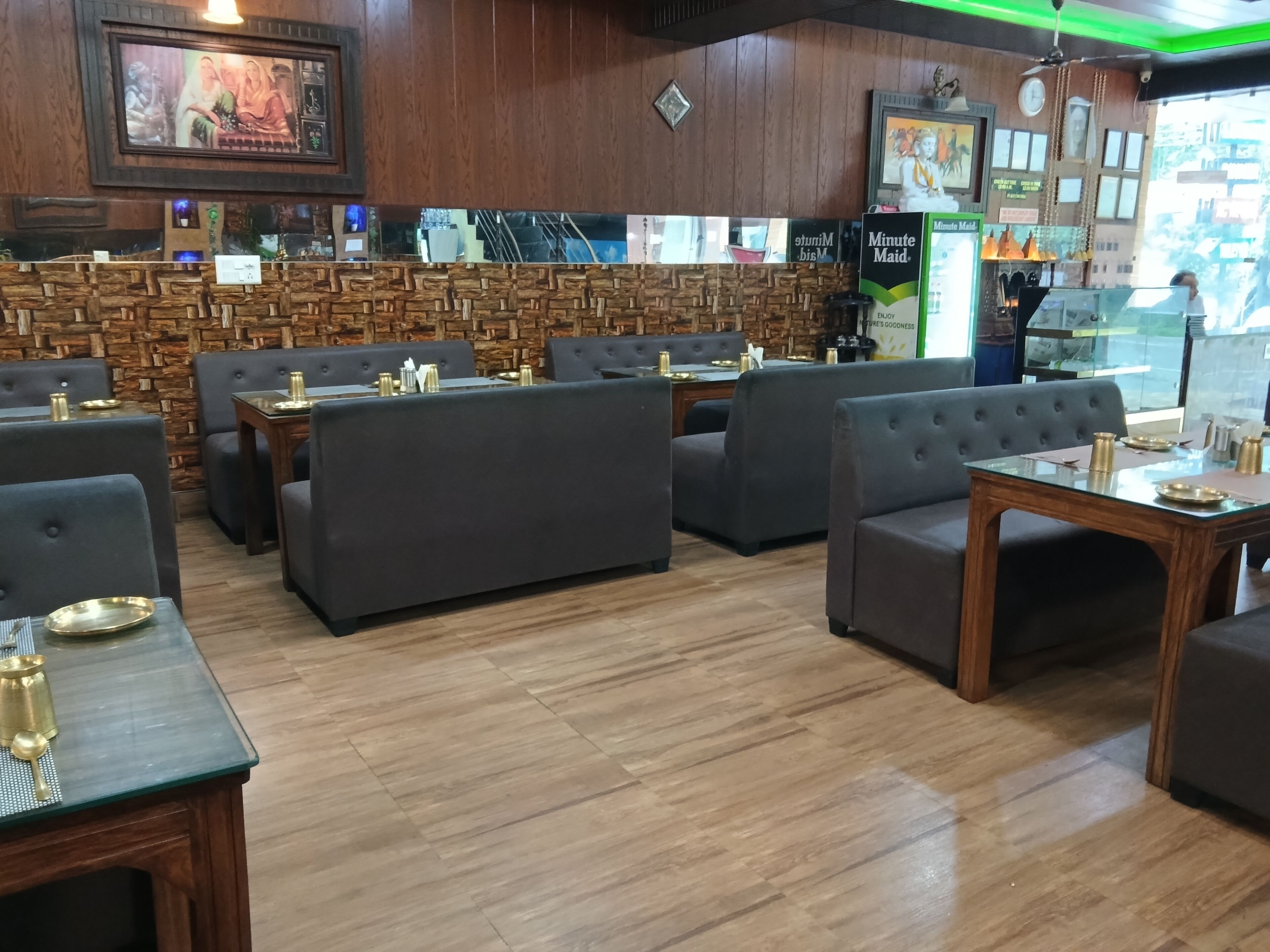 A restaurant interior with wooden walls, a stone accent wall, and a green-lit ceiling. Tables are set with brass plates and cups.