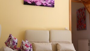 A room with a beige sofa, a floral-patterned pillow, and a wall-mounted painting of purple flowers. - Ferienwohnung in Freital, Dresden (Freital)