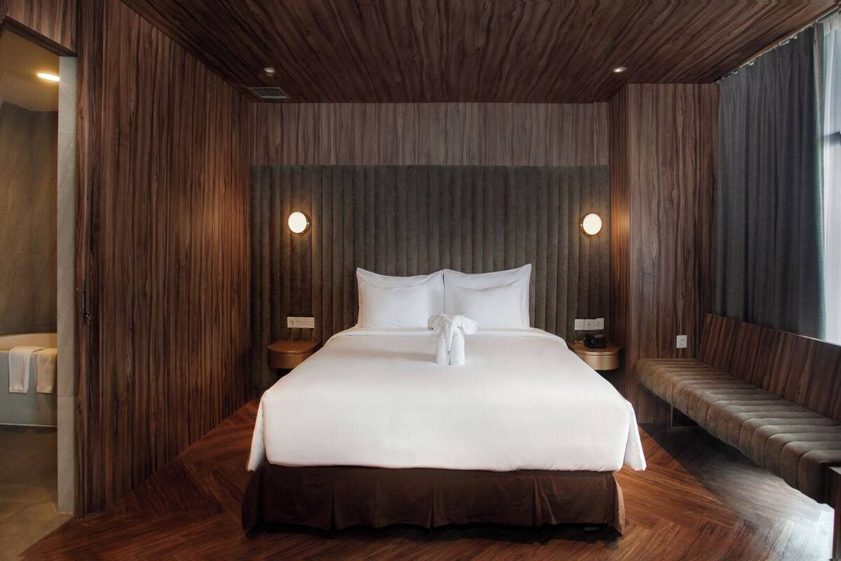 A modern hotel room with a large bed, wooden panel walls, and a bench by the window.