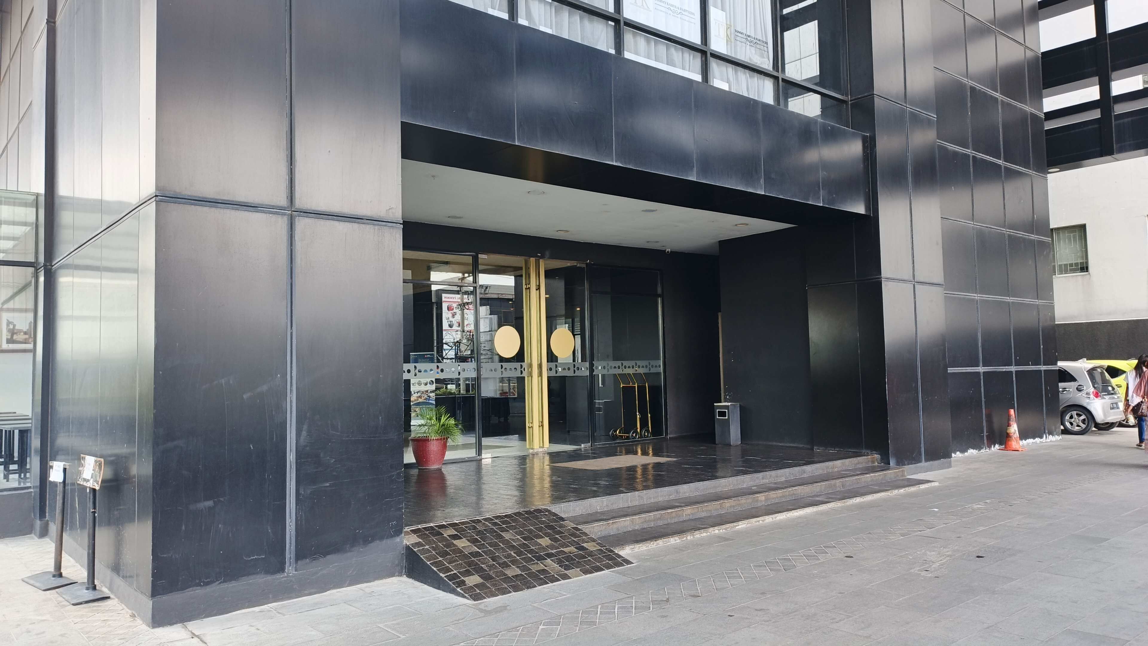 A modern building entrance with a glass door, a small outdoor seating area with plants, and a visible signboard.