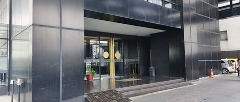 A modern building entrance with a glass door, a small outdoor seating area with plants, and a visible signboard.