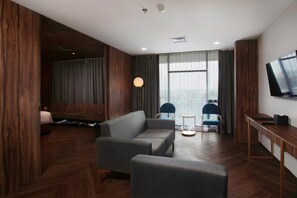 A modern hotel room with a gray sofa, two chairs, a TV, and a wooden accent wall. - Bono Hotel by ARTOTEL - Pekanbaru (Pekanbaru)
