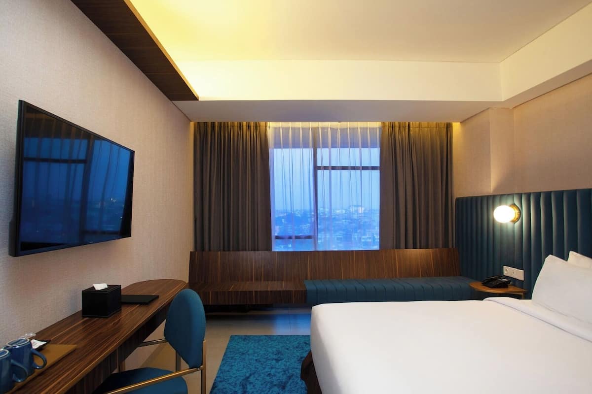 A hotel room with a bed, a desk, a chair, a TV, a window with curtains, and a nightstand with a lamp.