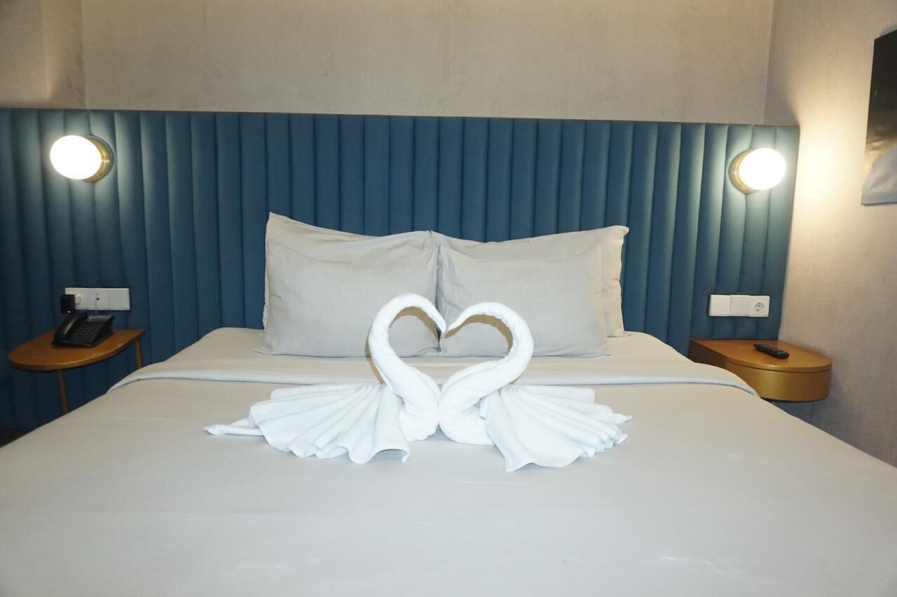 Ein Hotelzimmer mit Bett, Kissen, einer Handtuch-Herz-Arrangement, Nachttischen und Wandleuchten.