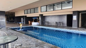 Une piscine intérieure avec des bords carrelés, un espace détente avec des tables et des chaises, et une vue sur la pièce adjacente grâce à un mur en verre.