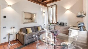 A modern living room with a glass dining table, a sofa, and a kitchen area with a microwave and kettle.