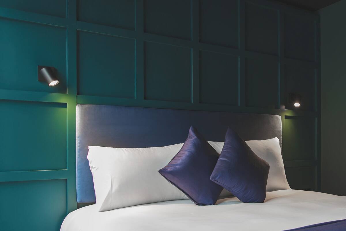 A neatly made bed with white and dark blue bedding, a dark headboard, and two wall-mounted lights.