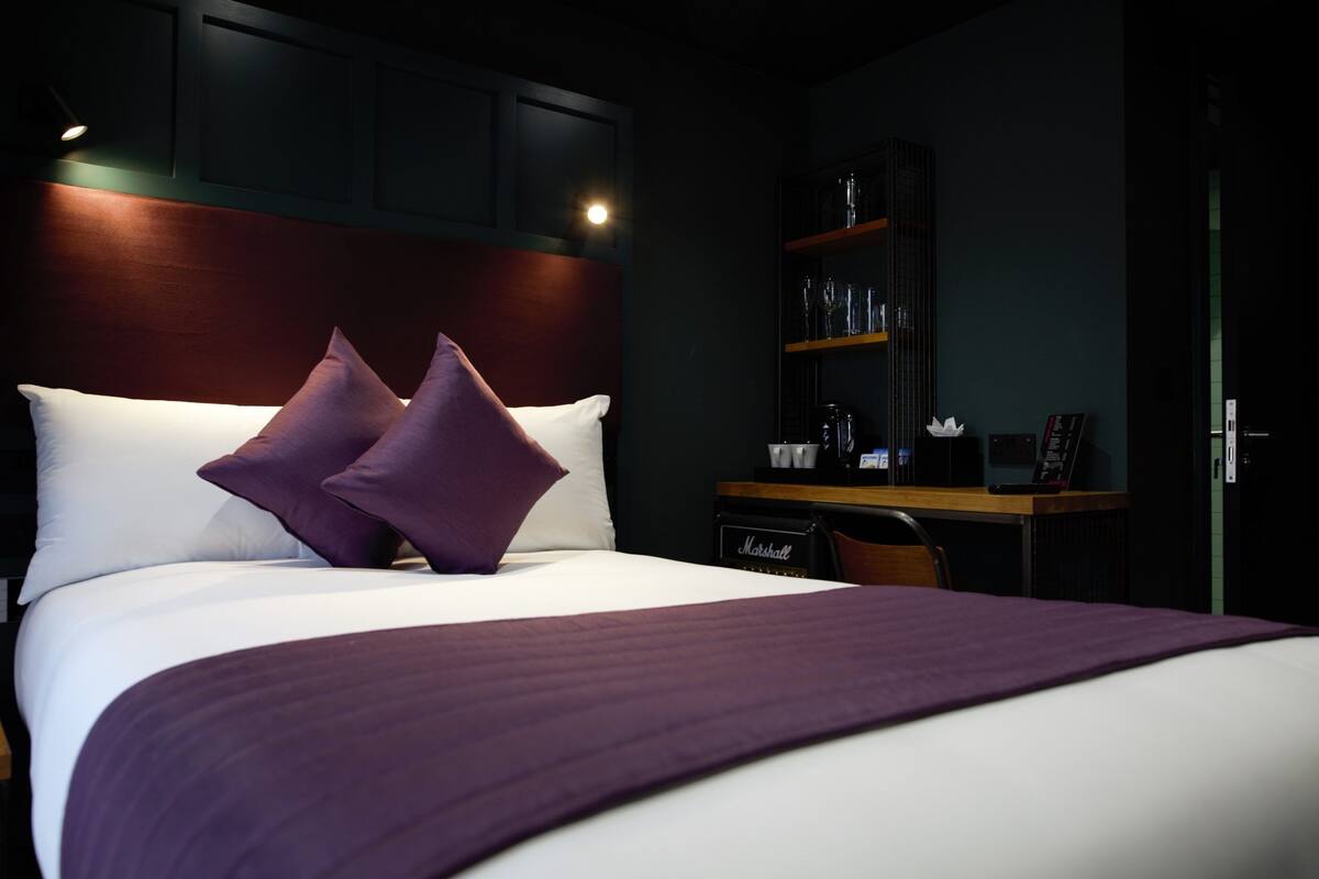 A hotel room with a large bed, two pillows, a dark headboard, a desk with a computer, and a shelf with glasses and a phone.