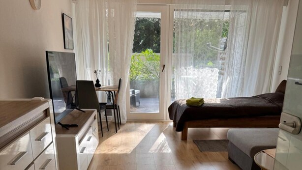 Ein kompaktes Hotelzimmer mit Küchenzeile, einem Bett, einem Schreibtisch mit Stuhl und einer Glasschiebetür, die auf einen Balkon führt.