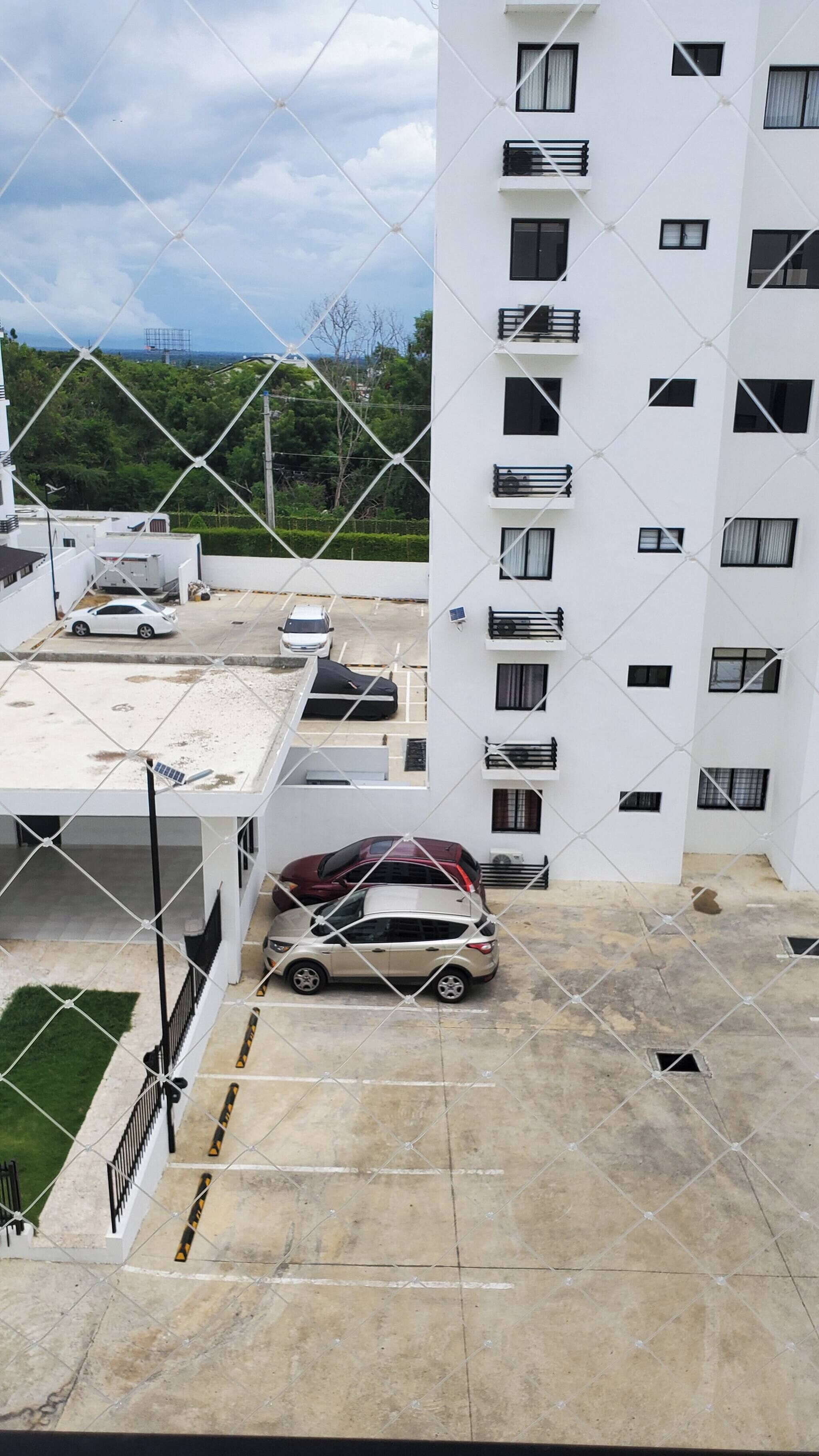 A multi-story building with a white exterior, multiple balconies, and a parking area with several cars.