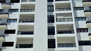 A multi-story building with a central vertical structure, multiple balconies, and a parking area in front. - 2BR 2BA Backup Power AC Wifi Gym 24h Security Ideal for Long Stays (Santiago de los Caballeros)