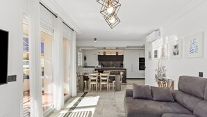 Een moderne woonkamer met een grijze bank, een glazen salontafel en een keuken op de achtergrond.
