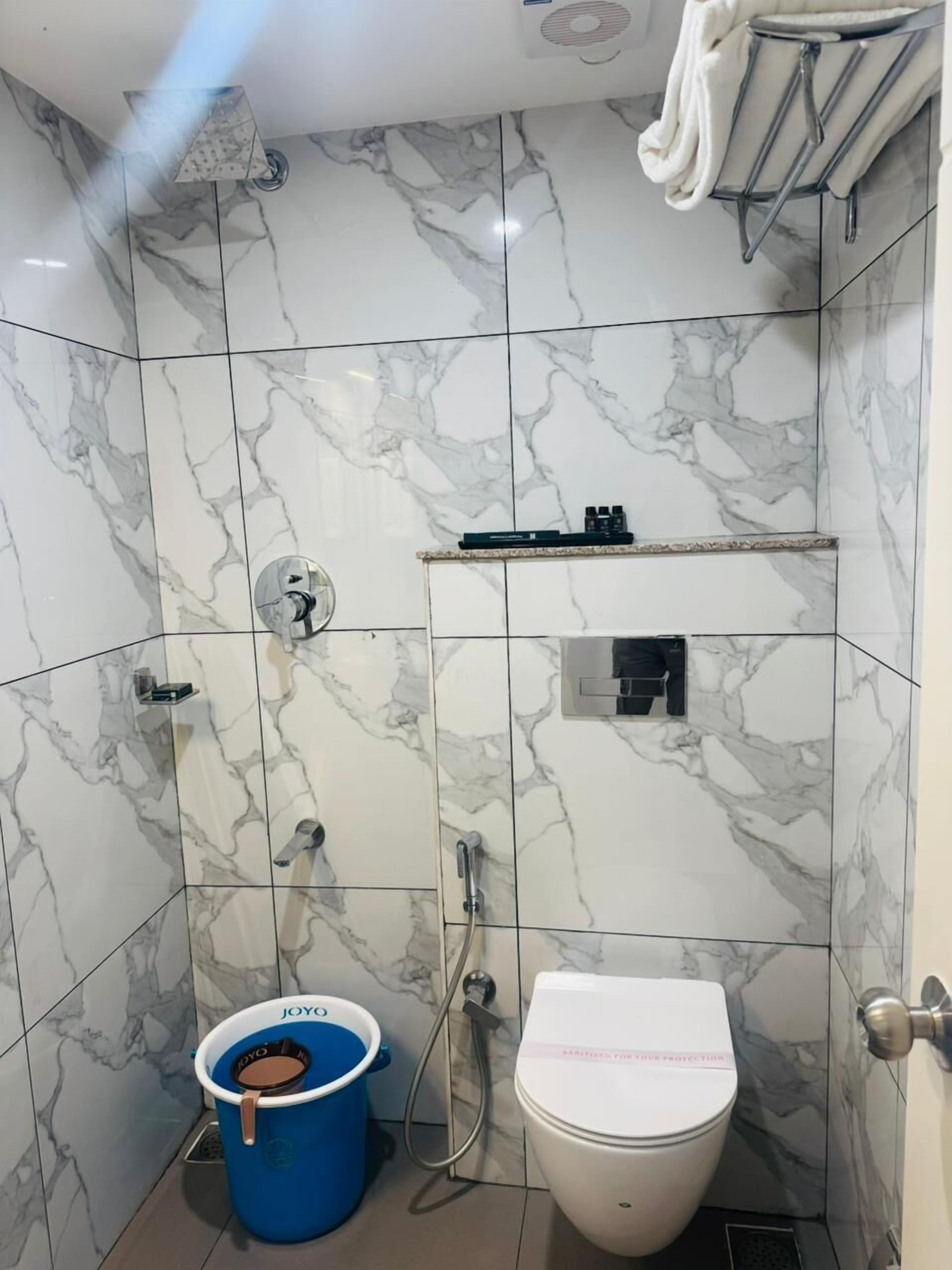 Un baño con azulejos de mármol blanco, un inodoro, una ducha, un cubo azul y un perchero para toallas.