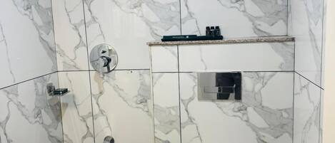 Un baño con azulejos de mármol blanco, un inodoro, una ducha, un cubo azul y un perchero para toallas.