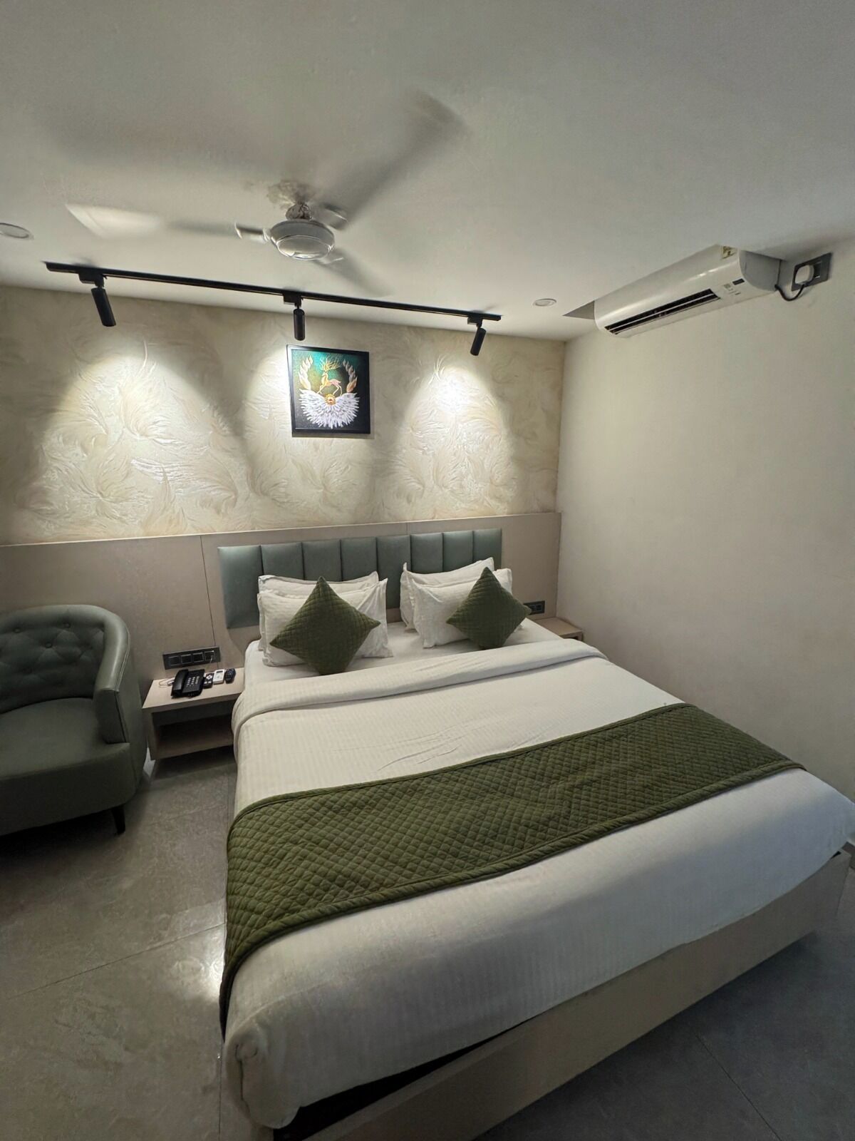 Habitación de hotel con cama, una silla, mesita de noche, ventilador de techo, equipo de aire acondicionado y una obra de arte colgada en la pared.