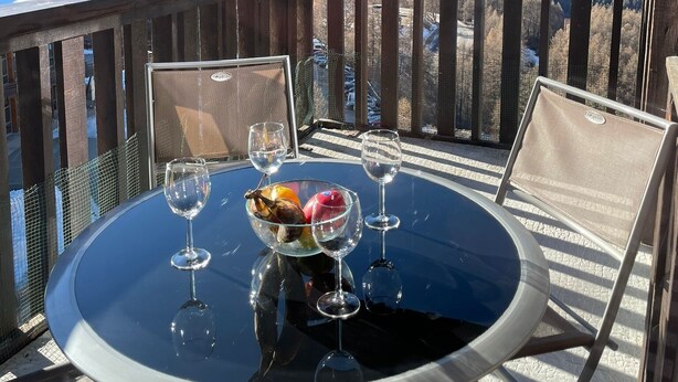 A round table with a glass top, two wine glasses, and a bowl of fruit on a balcony with a mountain view.