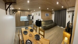 A modern kitchen with wooden cabinets, a dining area with a wooden table set for four, and a grey sofa in a living area.