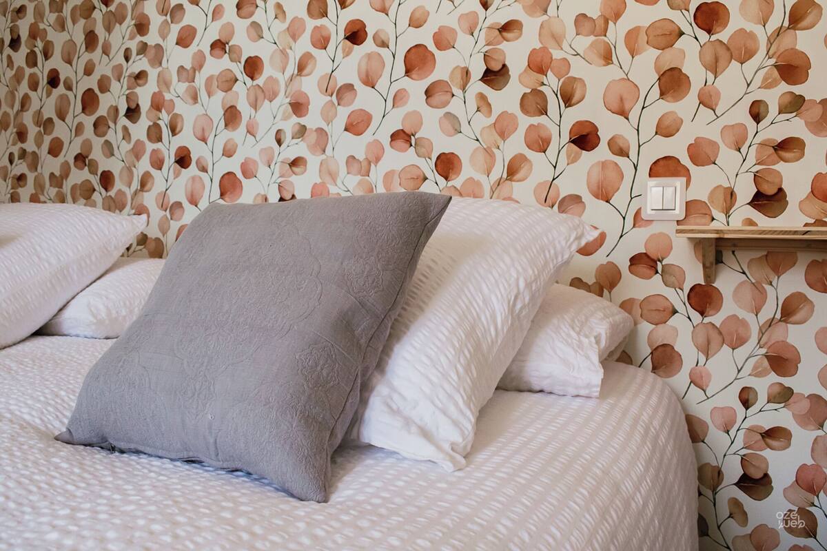 Ein ordentlich bezogenes Bett mit einem grauen Zierkissen und weißen Kissen, vor einer Wand mit rahmengemustertem Tapeten.