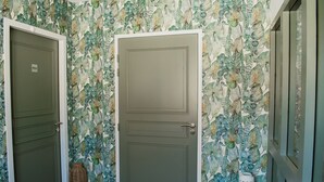 Un pasillo con dos puertas, una cerrada y otra abierta, que conduce a una habitación con papel tapiz estampado y una pequeña mesa con una maceta blanca y una taza.