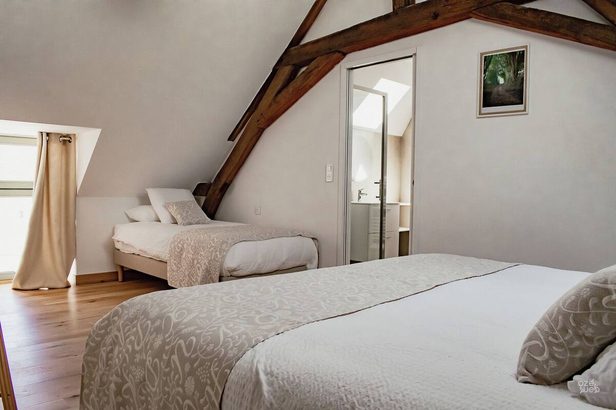 Ein Schlafzimmer mit schrägverlaufender Decke, einem großen Bett, einem kleineren Bett und einem durch eine offene Tür einsehbaren Badezimmer.