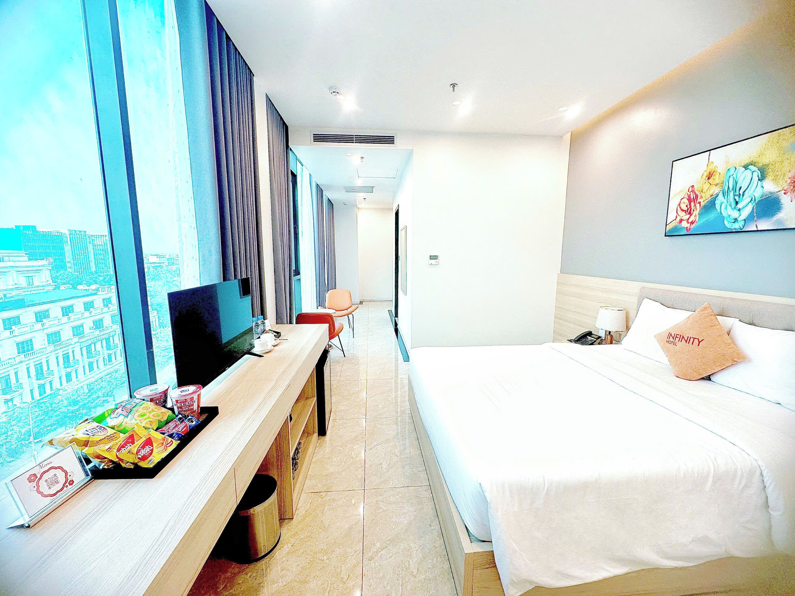 A hotel room with a bed, a TV, a desk with snacks, and a painting on the wall.