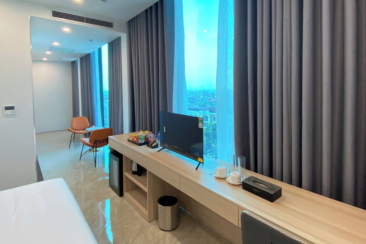 A modern hotel room with a large bed, a desk with a computer, and a view of the city through large windows.
