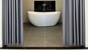 Une salle de bain moderne avec une baignoire sur pieds, une pomme de douche fixée au mur et des rideaux gris qui encadrent l’espace.