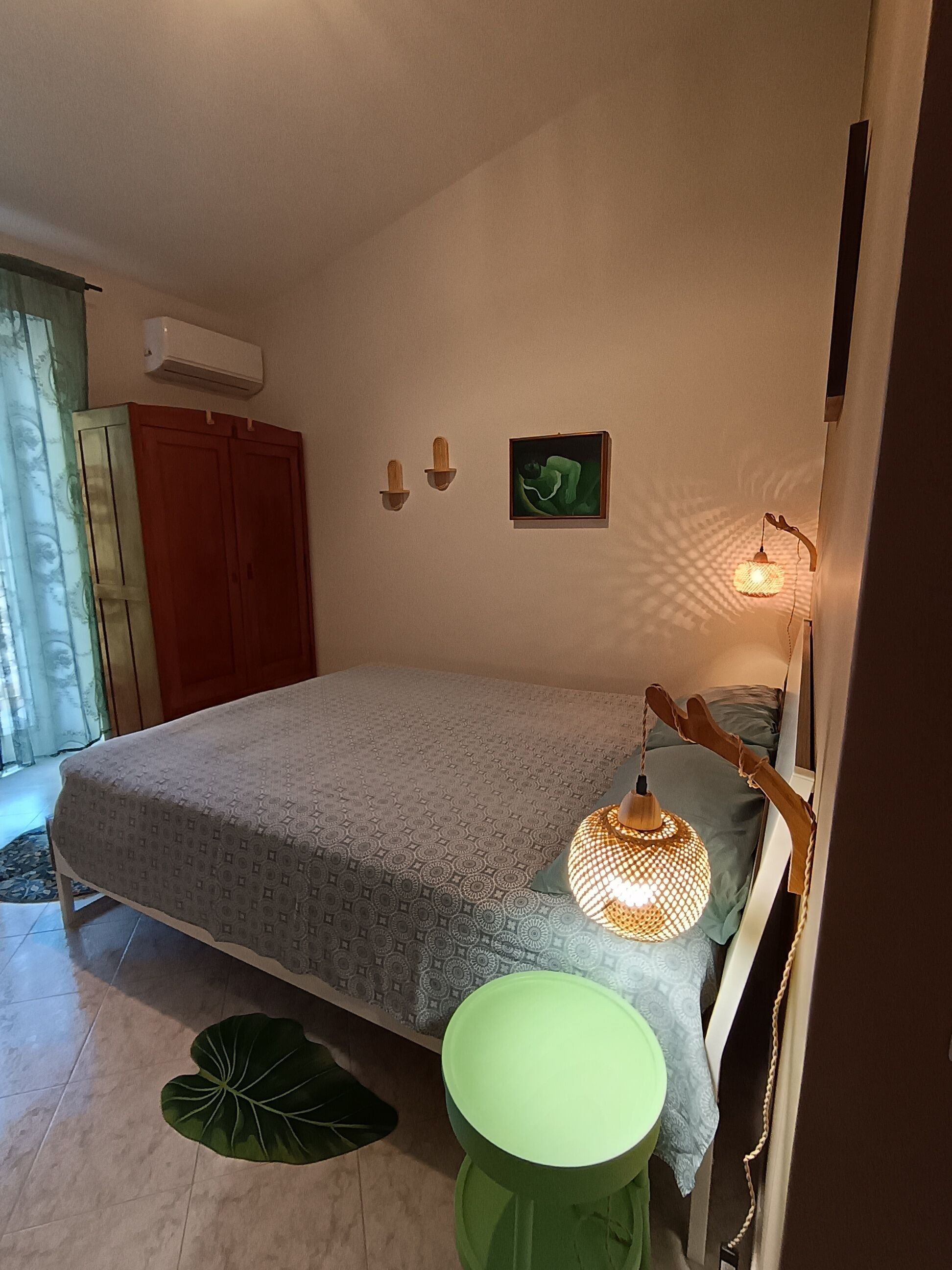 Une chambre à coucher avec un lit, un tabouret vert, une lampe fixée au mur et un tableau.