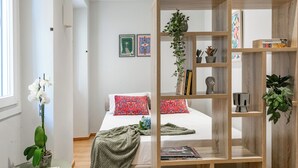 Une chambre à coucher avec un lit, une bibliothèque en bois, une plante en pot et une petite table sur laquelle se trouve un livre.