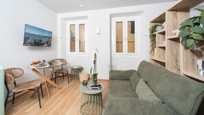 Ein modernes Wohnzimmer mit einem Esstisch aus Glas, Korbstühlen, einem grünen Sofa und einem hölzernen Bücherregal.
