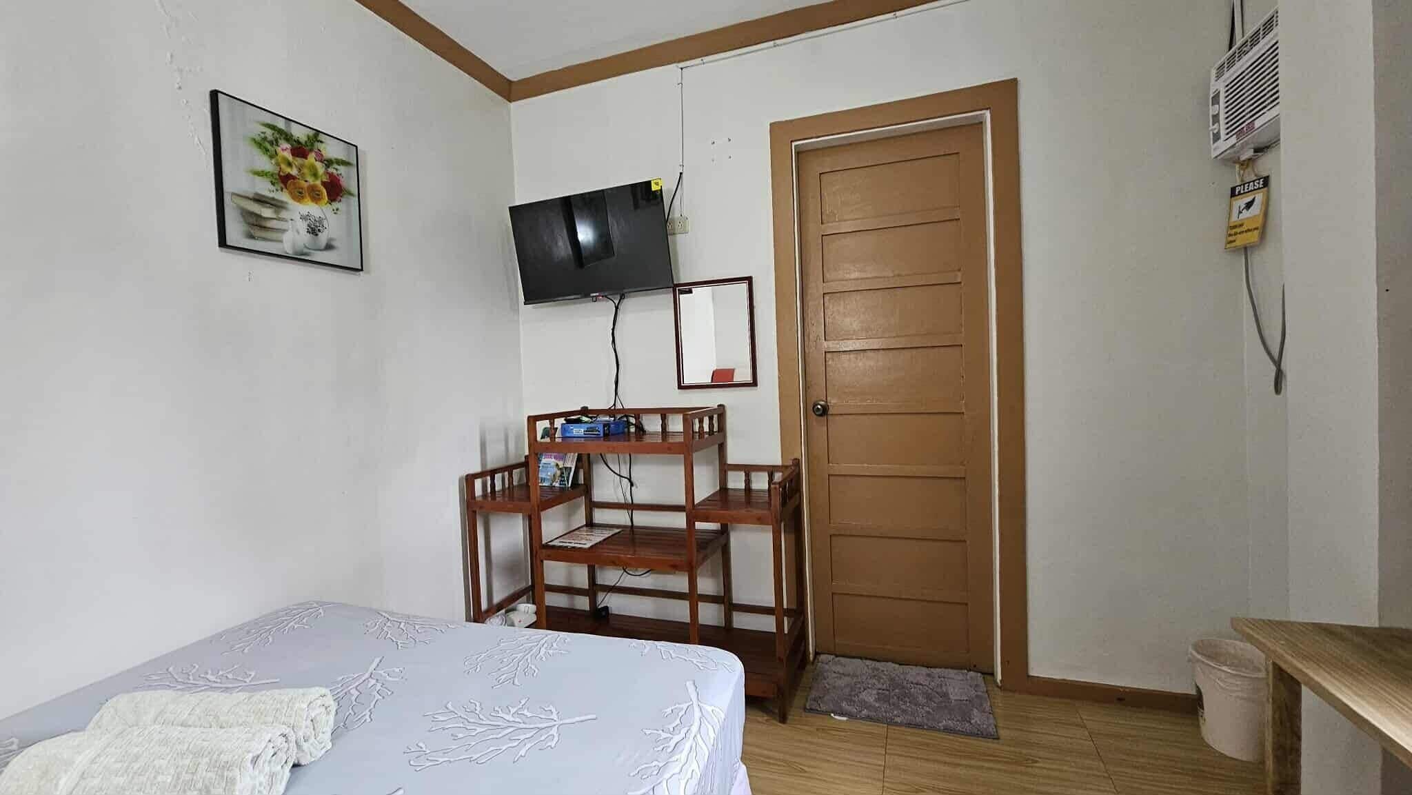 A small, simple room with a bed, a desk, a chair, a television, and a door.