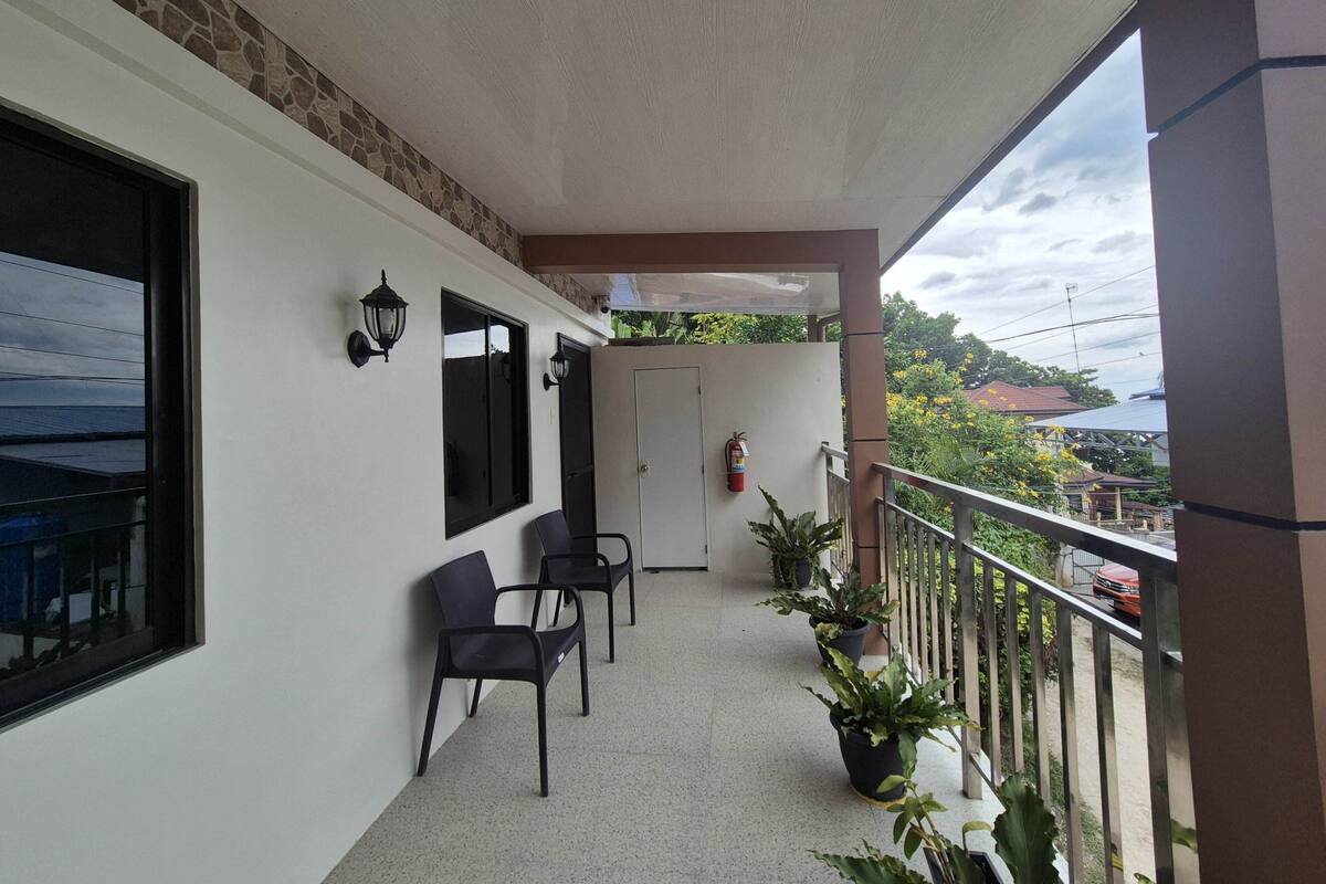 Un balcon avec un mur blanc, deux chaises noires, des plantes en pot, une porte et un extincteur.