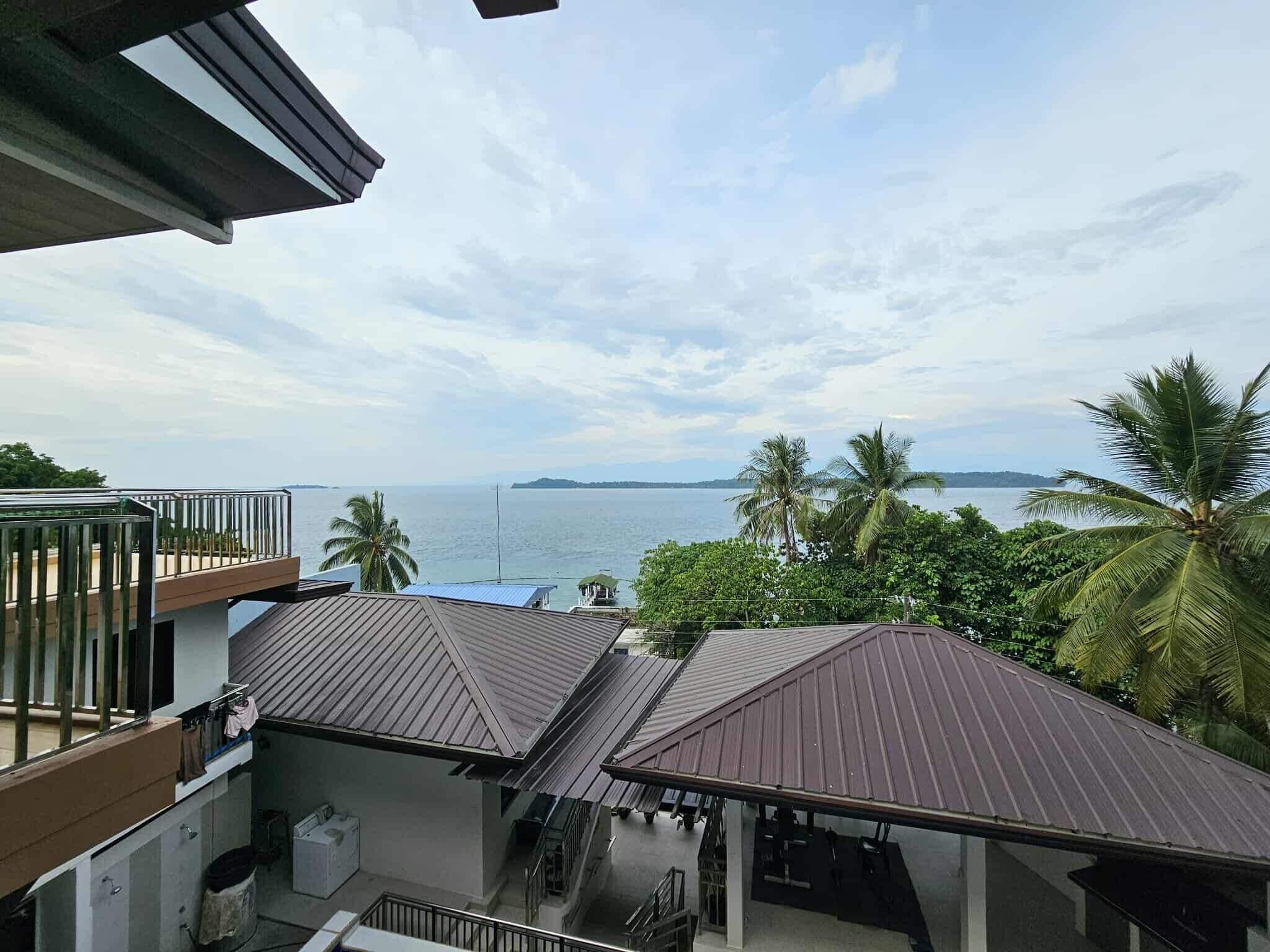 A view of a coastal property with a balcony overlooking the sea, a covered outdoor area, and a staircase leading to an upper level.