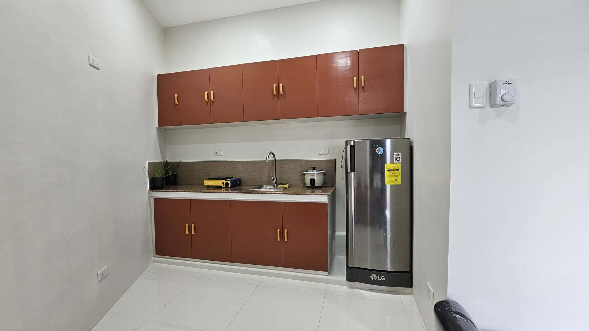 Cocina compacta con armarios marrones, un refrigerador de acero inoxidable y una encimera con hervidor y olla de cocción lenta.