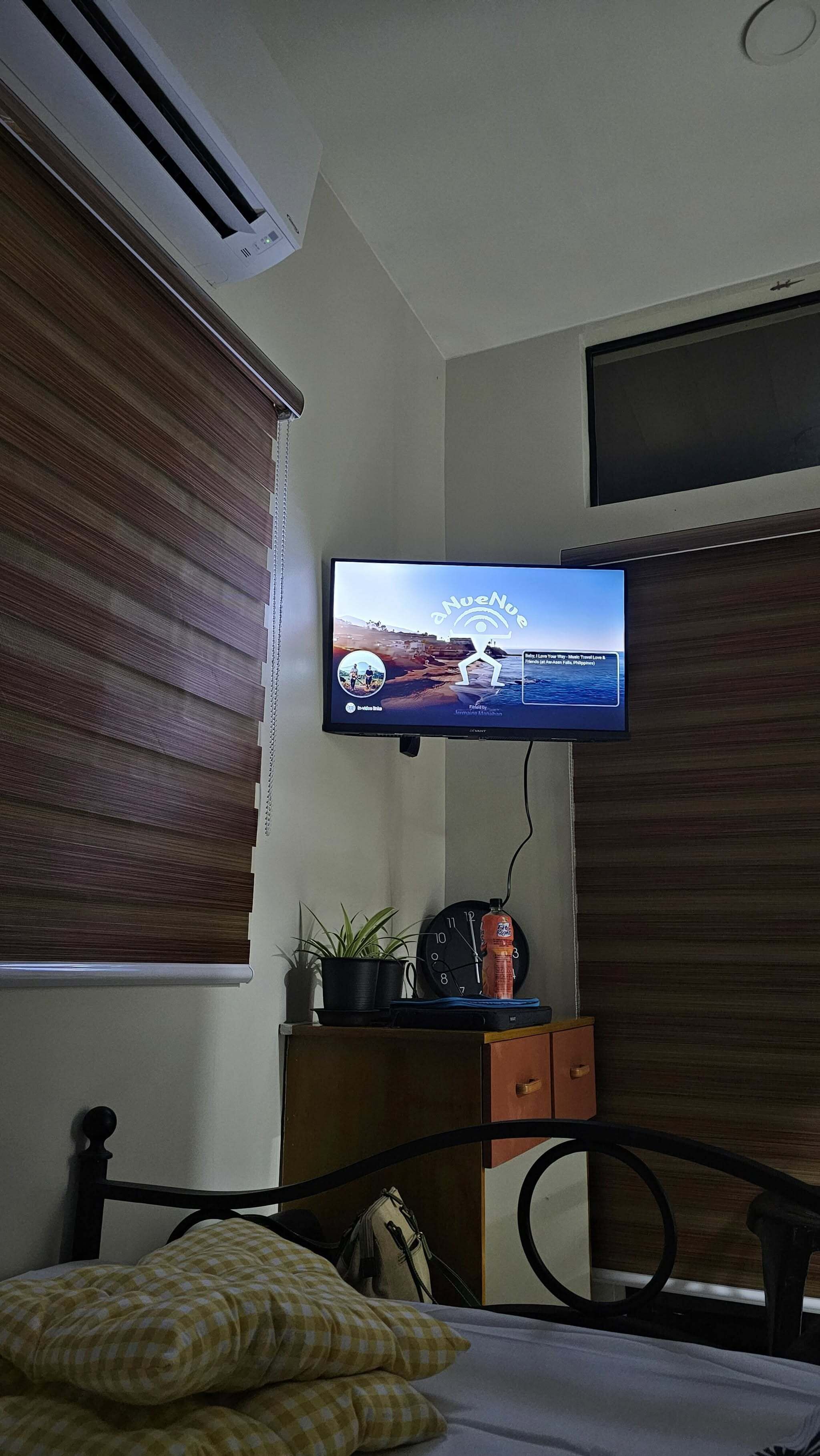 A hotel room with a television mounted on the wall, a bed with checkered bedding, a small table with a plant and a bottle, and a clock.