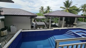 Ein Swimmingpool mit blauen Fliesen, ein überdachter Außenbereich mit Grill und im Hintergrund Palmen.