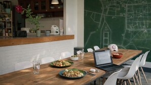 A dining area with a wooden table, white chairs, and a laptop. There are plates of food, a bowl, and a water glass on the table. A chalkboard with a map is in the background. - Adorable sunny bedroom with AC in Doi Suthep and nimmamhaemin neighborhood. (Muang Chiang mai)