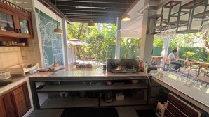 A kitchen with a gas stove, a sink, and a countertop with various items. There is a window with a view of greenery outside. - Adorable sunny bedroom with AC in Doi Suthep and nimmamhaemin neighborhood. (Muang Chiang mai)