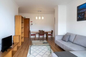 A modern living room with a grey sofa, a TV on a wooden stand, a dining area with a table and chairs, and a staircase leading to an upper floor. - Ocean View Balcony Apartment in Garajau, Madeira  Perfect R (Caniço)