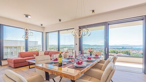 A modern dining area with a large table set for six, large windows offering a city view, and a red sofa in the background. - Aliara Panorama 1 
Luxusvilla (Vodice)